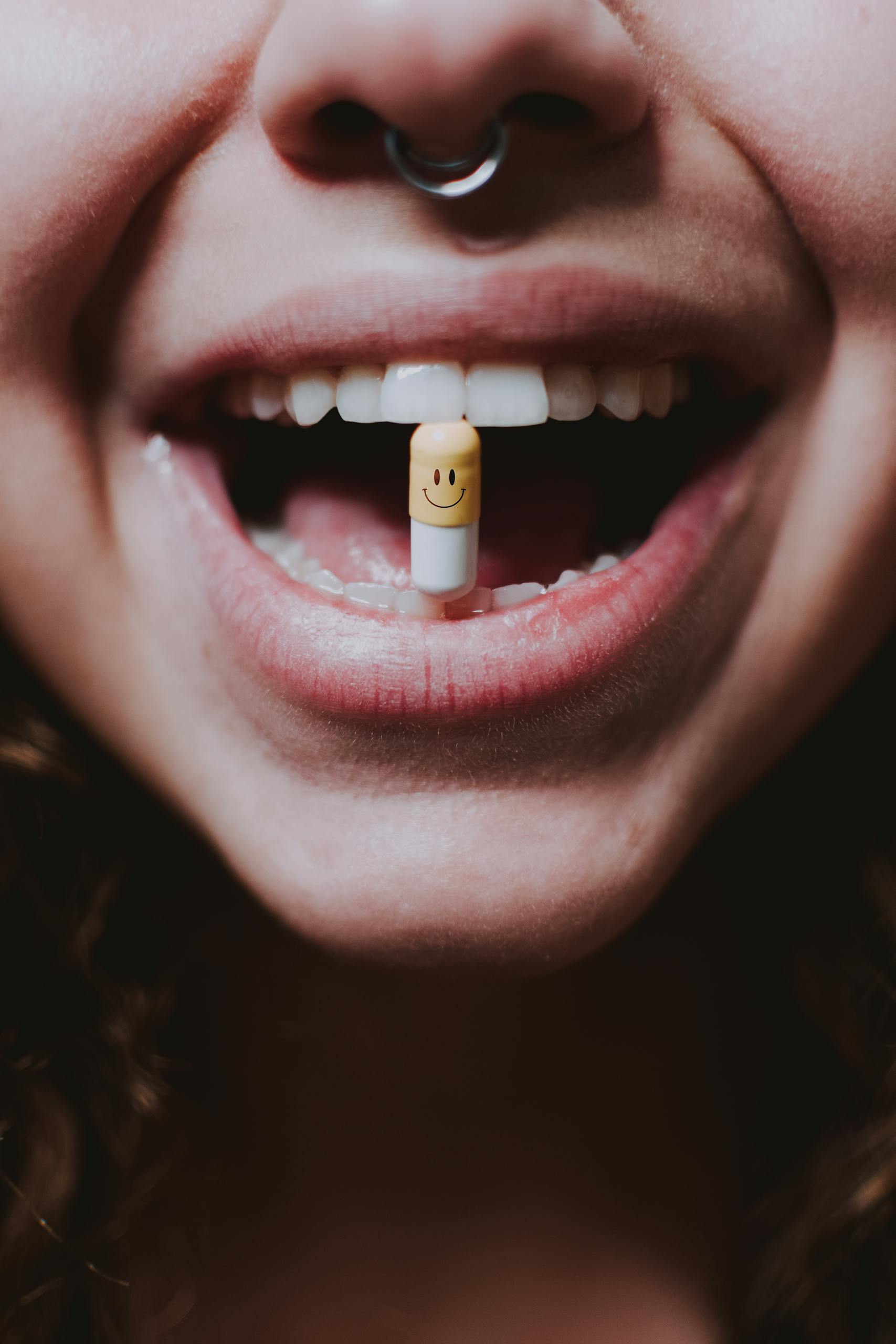 Close-up photo of a woman with a smiley face pill held between her teeth, evoking themes of medication and wellness.