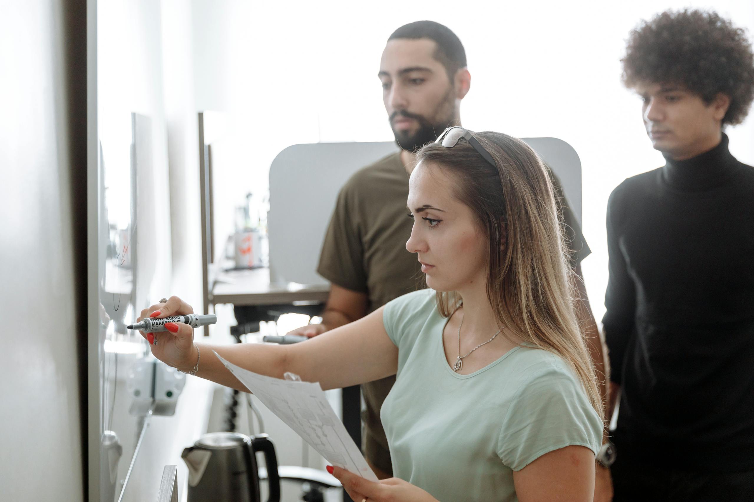 Three young professionals brainstorming on a whiteboard in a modern office setting.